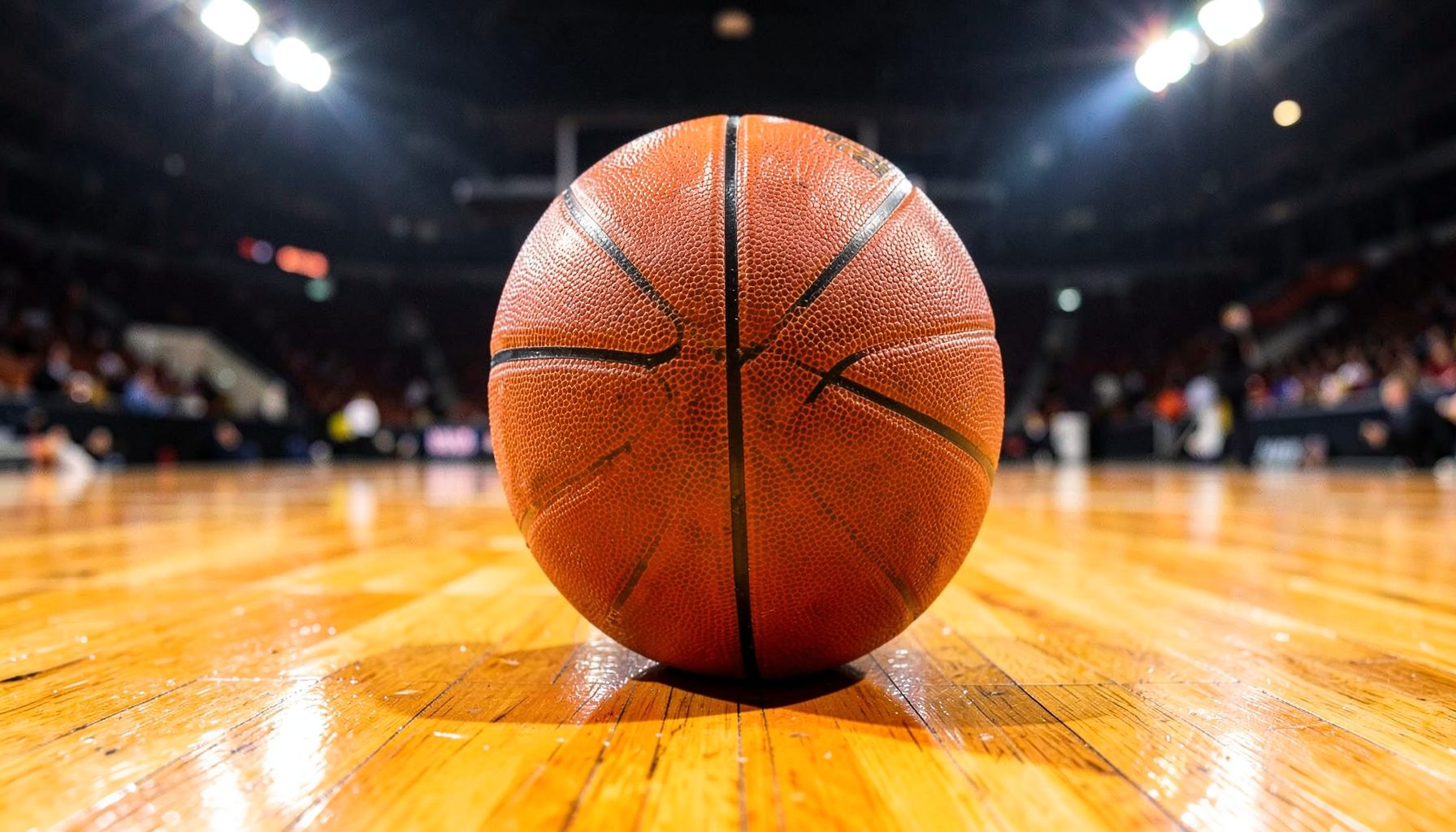 A basketball resting on a polished hardwood court under bright arena lights, warm wood tones and glossy orange.