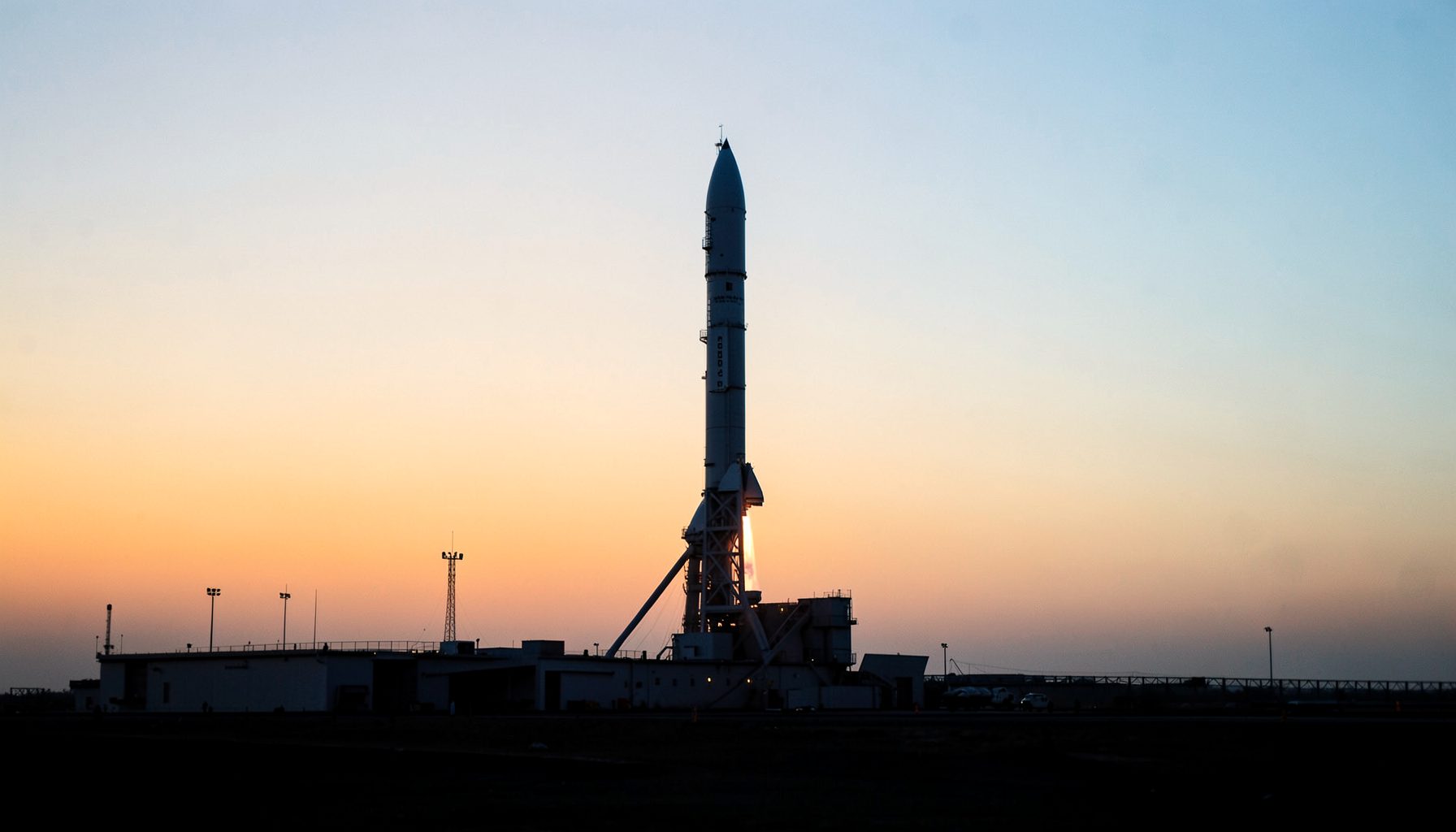A white rocket standing on its launch pad at sunset with a glowing orange sky and engine ignition visible