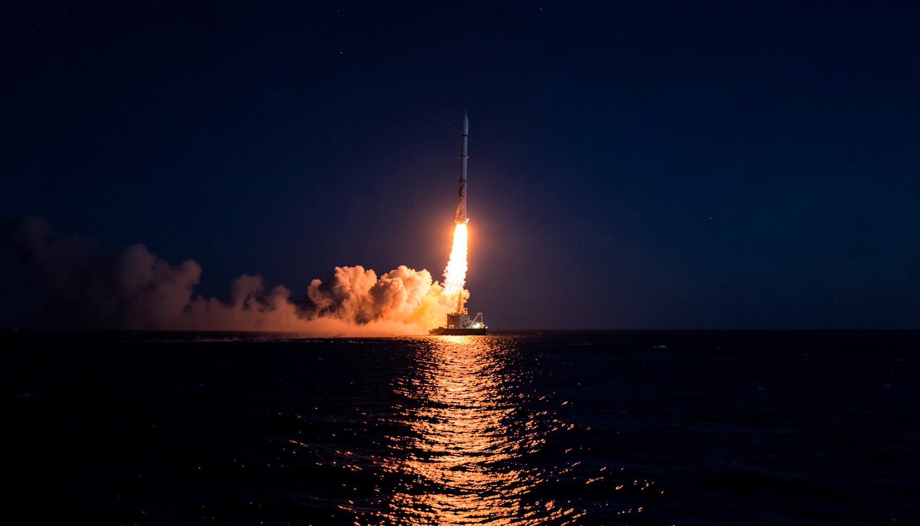 Nighttime rocket launch over dark ocean water, bright orange flame and smoke reflecting on the sea