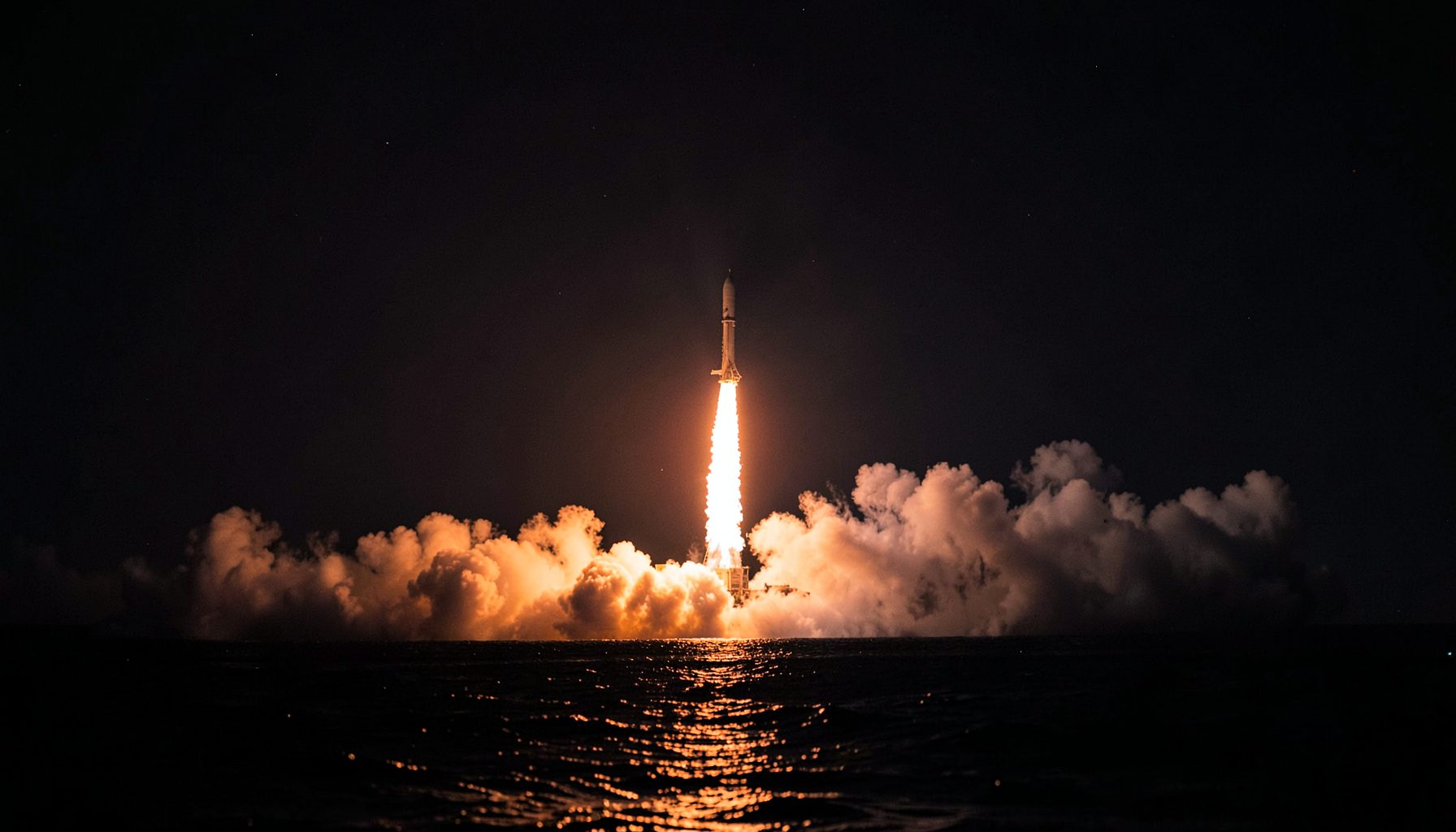 A rocket launching at night over the ocean, bright flames and smoke reflecting on dark water under a starry sky.