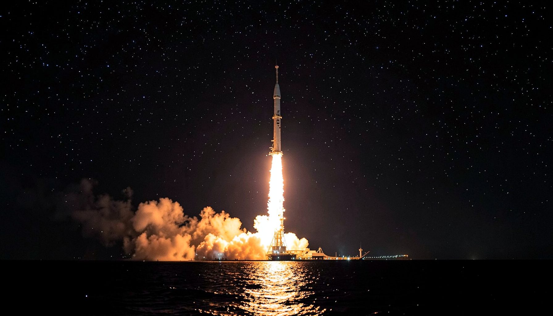 A rocket launching at night from a seaside platform with bright flames, smoke, and a starry sky reflected on the water