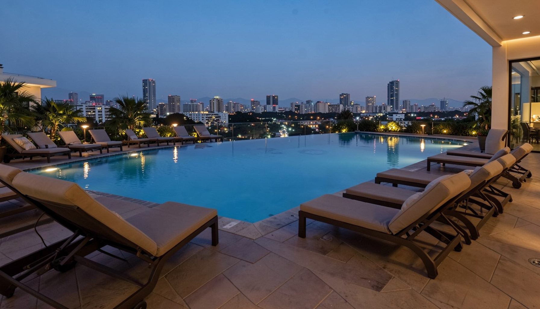 An infinity pool with lounge chairs on a terrace overlooking a lit city skyline at dusk, blue and warm-toned lighting
